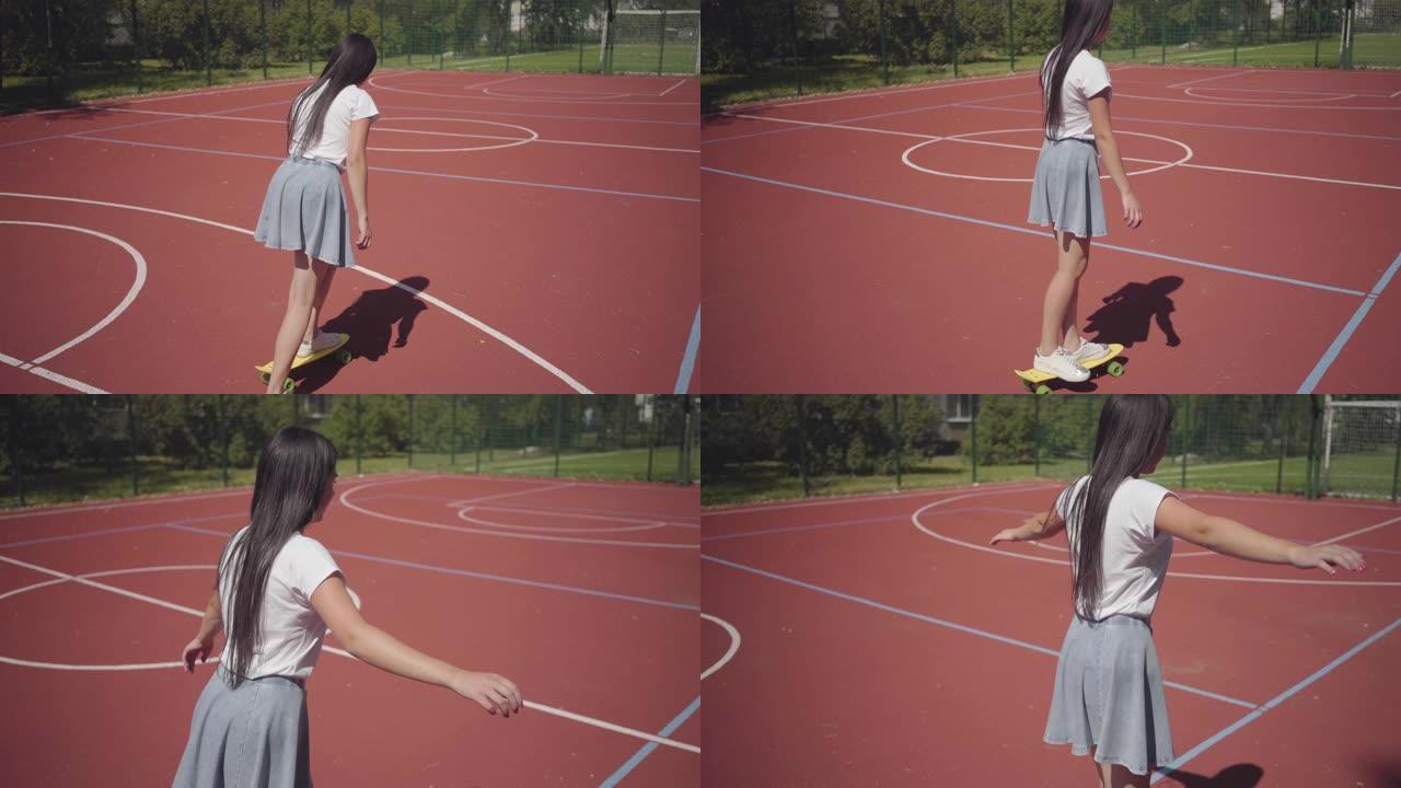 穿着运动鞋、裙子和t恤的少女在室外篮球场上骑着黄色滑板。