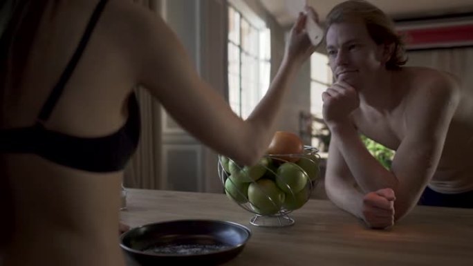 年轻的裸体夫妇站在厨房桌子旁，穿着内衣的性感女孩在煎锅里混合盐，用勺子敲打她穿着内衣的男人。男人向后