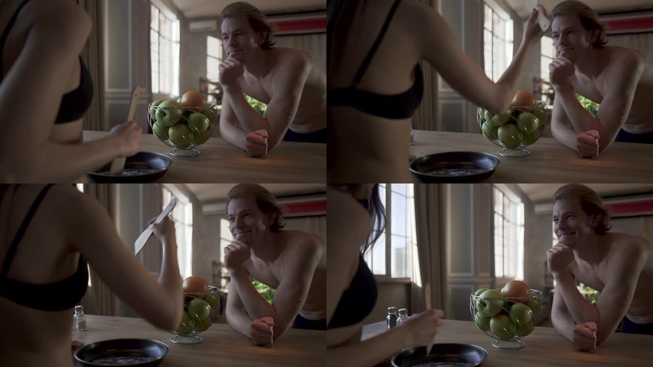 年轻的裸体夫妇站在厨房桌子旁，穿着内衣的性感女孩在煎锅里混合盐，用勺子敲打她穿着内衣的男人。男人向后
