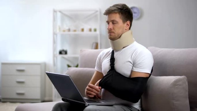 在笔记本电脑上工作的泡沫颈圈和手臂吊带受伤的男子感到疼痛