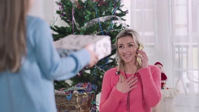 迷人的白人妇女在女儿带来圣诞礼物时收起智能手机。惊讶的女人在除夕夜收到黑发可爱女孩的礼物。电影院4k
