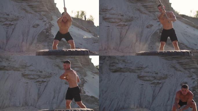 日落时分，在沙质山脉中，肌肉运动员在一个巨大的轮子上用慢镜头敲击锤子。车轮扬起尘土。