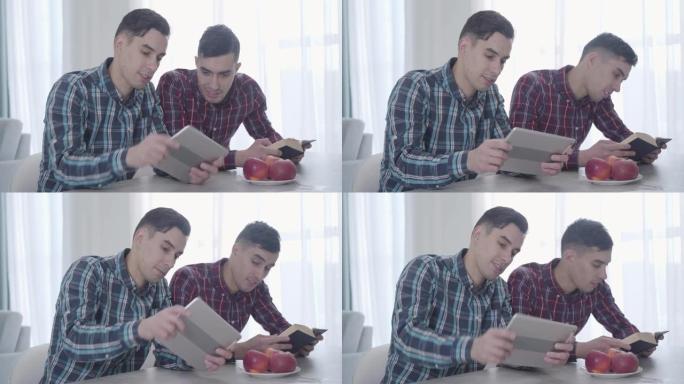 成年白人男子在平板电脑上玩电子游戏并展示给他的双胞胎兄弟的肖像。黑发男人看书，看着设备屏幕。兄弟姐妹