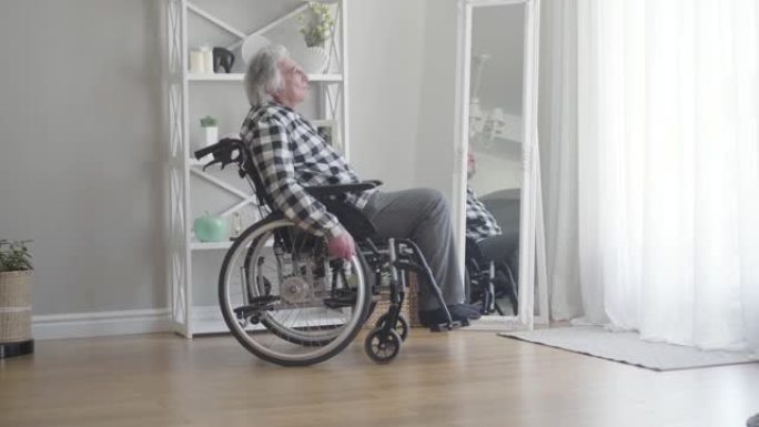 坐在轮椅上的白人老人在家滚动到窗户的侧视图。沮丧的老退休人员交叉双手，看着远处思考。悲伤，退休，孤独
