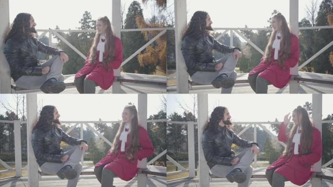 欢快的年轻混血儿夫妇坐在亭子的长椅上，有说有笑的肖像。积极嬉皮士男女在秋季公园约会。爱情，浪漫，幸福
