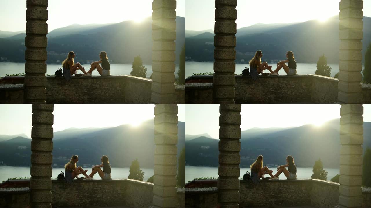 两名年轻妇女坐在石墙上，被柱子框住，俯瞰湖泊和山脉