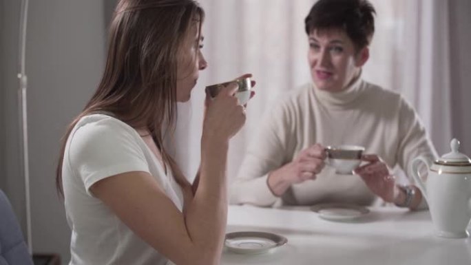 侧视特写照片，年轻的高加索女孩喝热茶，坐在背景上与母亲交谈。女儿在家享受和妈妈在一起的时光。专注于前