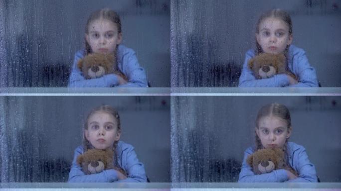 美丽的小女孩在下雨的窗户后面拥抱泰迪熊，欺负，孤独