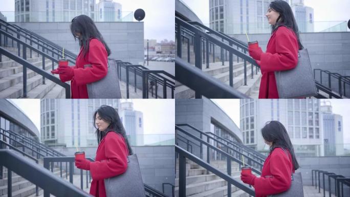 正面黑发高加索女孩在城市楼梯上的侧视图。穿着红色外套的年轻美女带着咖啡杯漫步。生活方式，城市生活，幸