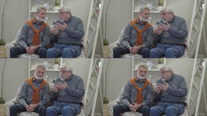 两名高加索老人在疗养院交谈的肖像。成熟的男性退休人员坐在沙发上聊天。