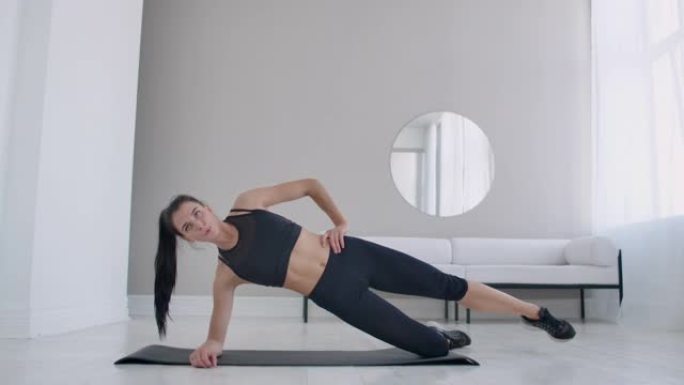 公寓里的黑发女人做一个锻炼的外侧木板站在她的膝盖上，抬起她的腿。健身马拉松