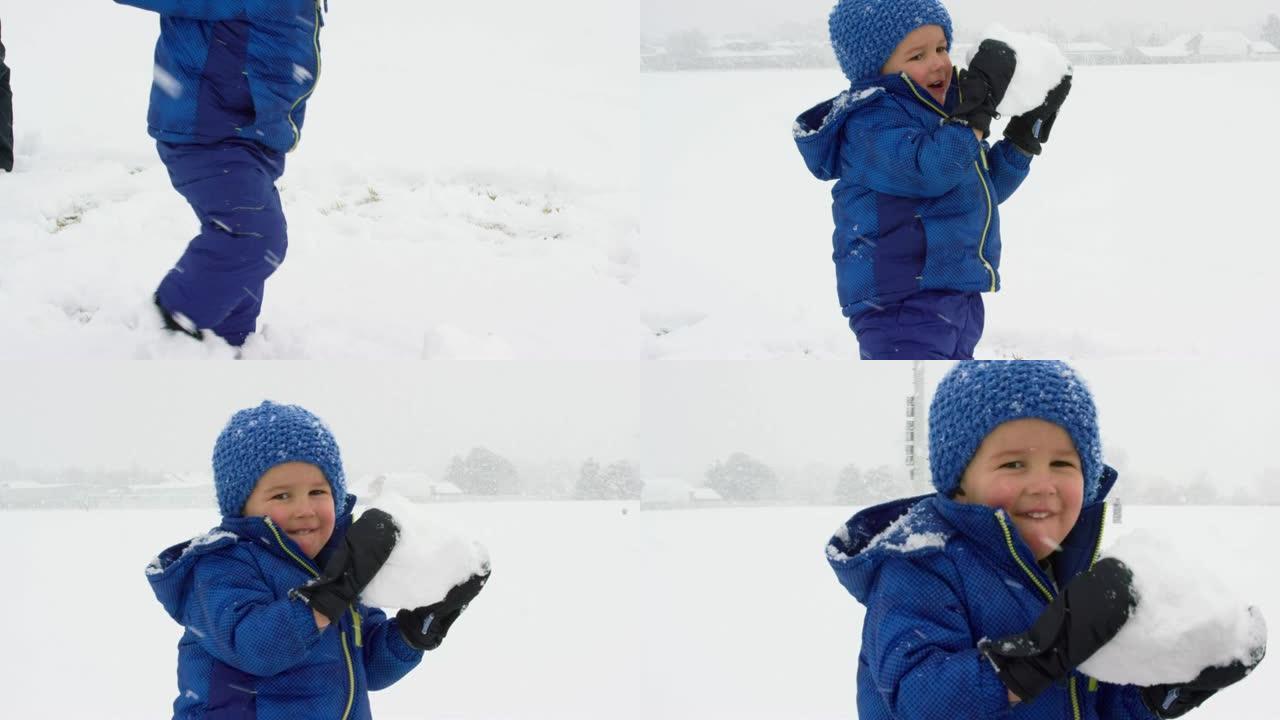 三岁的白人男孩穿着冬装在地上滚雪球，在下雪的阴天捡起来
