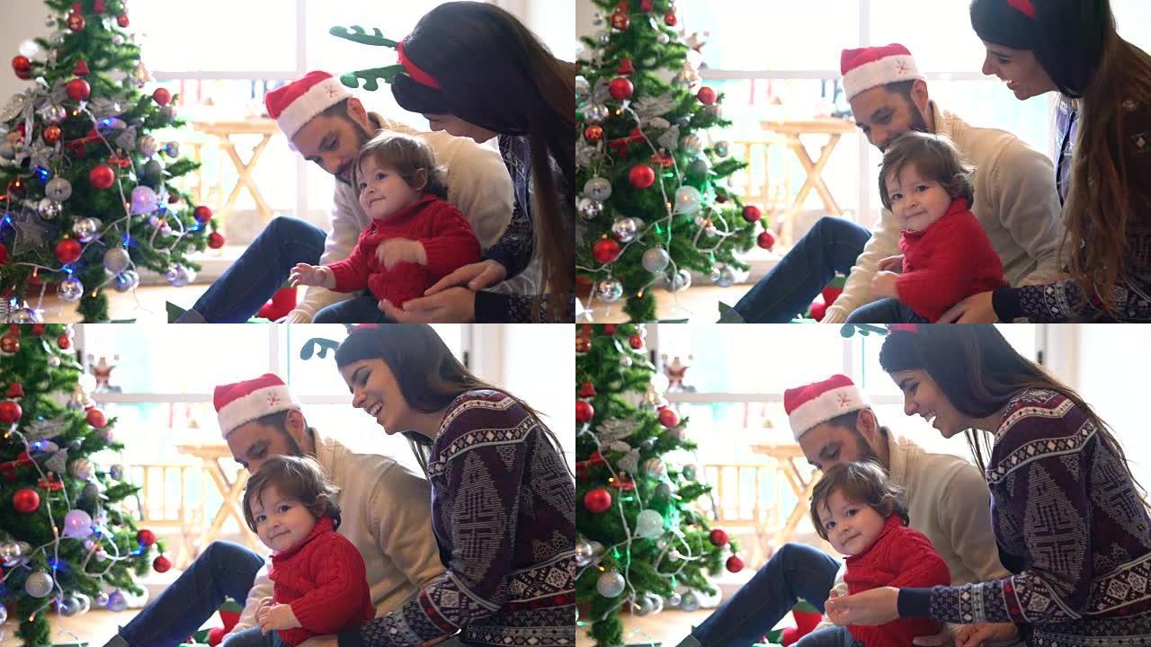 美丽的女婴在快乐的移动和鼓掌，而父母都微笑着坐在圣诞树旁边