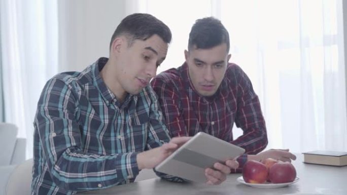 成年白人黑发男子阅读，作为他的双胞胎兄弟在平板电脑上玩游戏。那家伙放下书，看着设备屏幕。兄弟姐妹周末