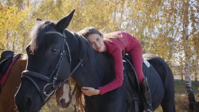 年轻的高加索女性马术者躺在马背上爱抚她的宠物。优雅的黑马站在秋天的森林中，年轻的骑手在他的背上。