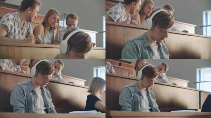大学礼堂的一名男学生在休息时戴着白色耳机听音乐，而不注意他人。观众中有很多人在谈论