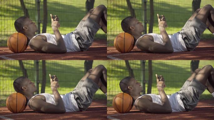 年轻的非洲裔美国人躺在球上并使用智能手机的侧视图。积极疲惫的篮球运动员在球场上休息和上网的肖像。休息