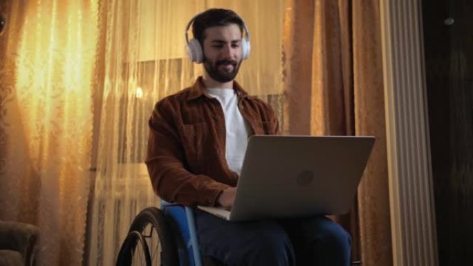 坐在轮椅上的严肃的年轻人使用笔记本电脑和耳机工作。残疾人在家里用笔记本电脑工作。