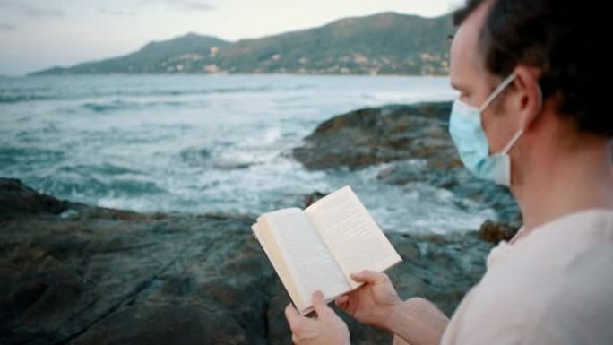 一个戴着面具的男人坐在海洋附近的岩石上看书的特写镜头