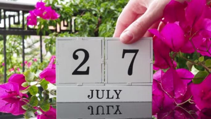 一个木制日历，有一个重要的活动，7月27日在一张有芙蓉花的桌子上，女人的手在日历上设定了日期。将日历