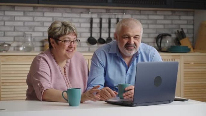 快乐的老年男人和女人正在用笔记本电脑与他们的朋友或家人在线聊天，在家庭厨房里肖像