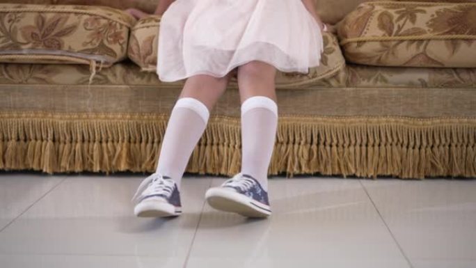 穿着衣服的白人小女孩的腿坐在室内客厅的沙发上。周末在家休息的无忧无虑快乐的孩子。生活方式和童年概念。