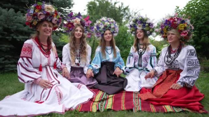 五名乌克兰年轻妇女坐在公园绿色的春夏草坪上唱民歌。美丽自信的积极女士的肖像，穿着民族服装和手持微笑的