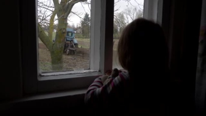 女童透过农村房的窗户看着拖拉机的工作