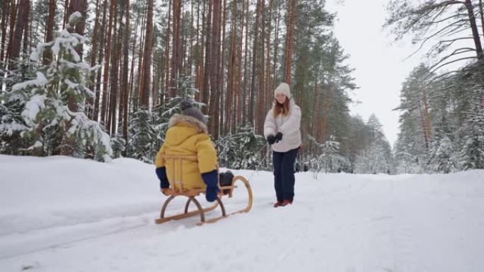 快乐的年轻女子和她的小孩在冬天的森林里散步，儿子坐在木制雪橇上