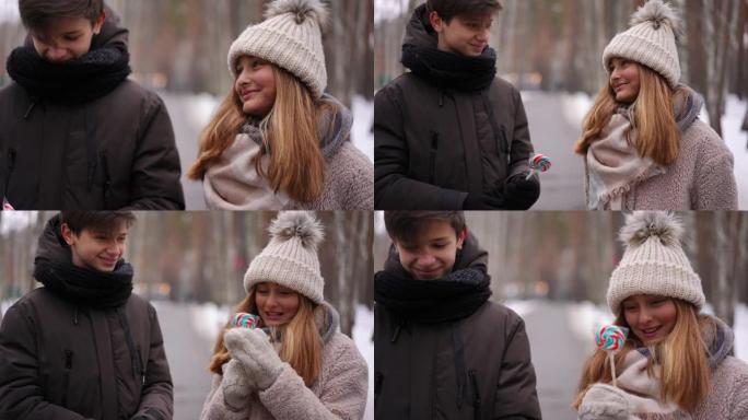 缩小放大微笑的少女在户外与男孩约会的肖像，男友以慢动作将棒棒糖传给女友。高加索青少年夫妇在冬季公园聊
