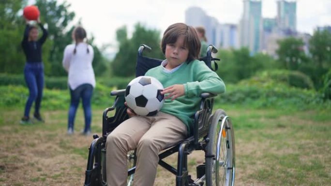 绝望的白人残疾男孩坐在轮椅上，拿着球看着相机，表情悲伤，朋友们在背景慢动作中玩耍。沮丧的孩子在公园摆