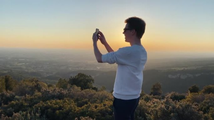 日落时站在山顶上的男子用智能手机拍照