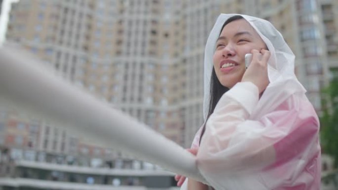 微笑的亚洲妇女在城市享受阴雨天气，在户外打电话
