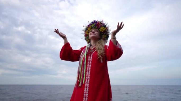 穿着红色连衣裙和花圈的微笑乌克兰妇女举起双手仰望多云的天空。穿着传统服装站在河岸欣赏自然的积极自信女