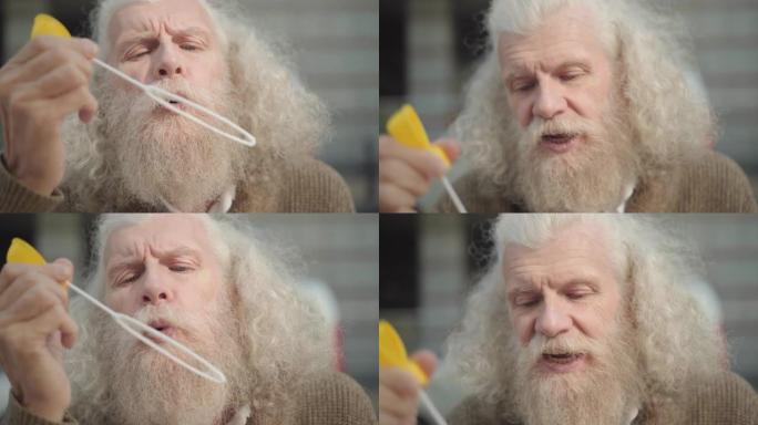 老人的特写脸，留着长长的白发和胡须，在户外吹肥皂泡。积极无忧无虑的男性白人退休人员在阳光明媚的日子享