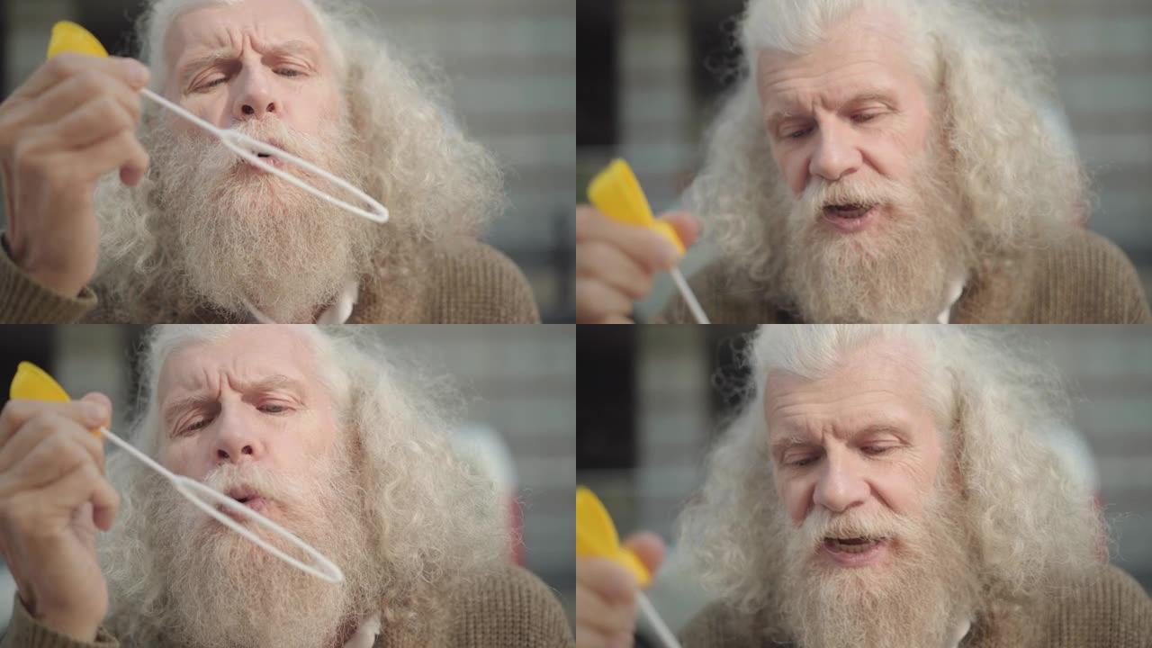 老人的特写脸，留着长长的白发和胡须，在户外吹肥皂泡。积极无忧无虑的男性白人退休人员在阳光明媚的日子享