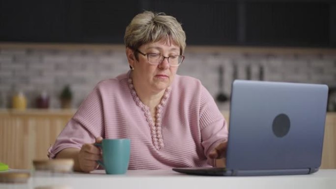 退休妇女正在上网，白天使用笔记本电脑坐在家里的厨房里，用现代笔记本的老太太肖像