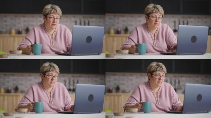 退休妇女正在上网，白天使用笔记本电脑坐在家里的厨房里，用现代笔记本的老太太肖像