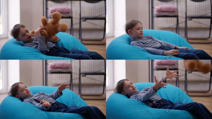 快乐的自闭症男孩躺在家里舒适的包椅上扔泰迪熊。笑高加索孩子的肖像，精神障碍在室内玩得开心。舒适和自闭