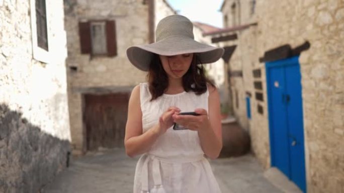 千禧一代高加索游客慢动作滚动智能手机屏幕，站在古镇户外环顾四周。年轻美女在塞浦路斯旅游观光的肖像。