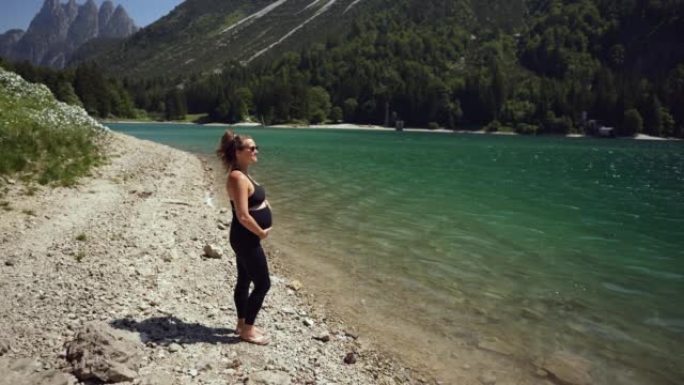 孕妇站在高山湖边望着群山，一边微笑着握住并揉着肚子