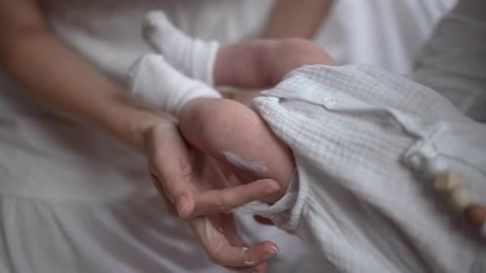 女性手在新生儿腿部慢动作涂抹润肤霜。无法辨认的高加索年轻母亲在家室内照顾婴儿男孩。