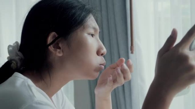 亚洲女孩通过智能手机观看电影或病毒视频，并在周末在家窗口附近吃零食。