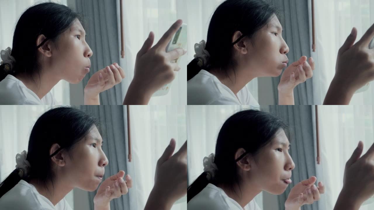 亚洲女孩通过智能手机观看电影或病毒视频，并在周末在家窗口附近吃零食。