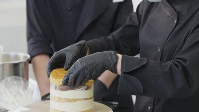 相机从女性的双手向上移动，将烘烤的圆形面团放在蛋饼上，到糖果人和受训者戴着Covid口罩的集中脸。厨