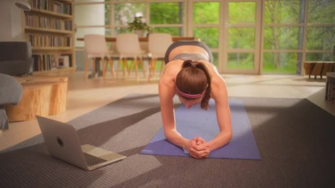 女运动员用笔记本电脑在瑜伽垫上做木板。放松的家庭锻炼