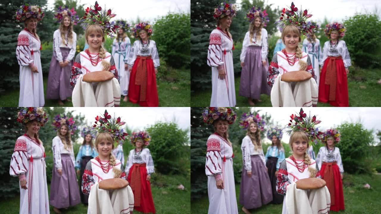 微笑的乌克兰小女孩站在毛巾上的传统面包上看着相机的肖像，背景模糊的女人。迷人自信可爱的孩子在公园户外