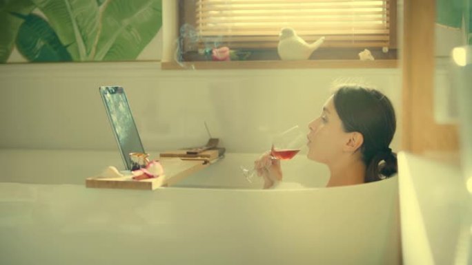 在浴室看电影。一个女人在浴缸里用笔记本电脑，喝着饮料吃着零食