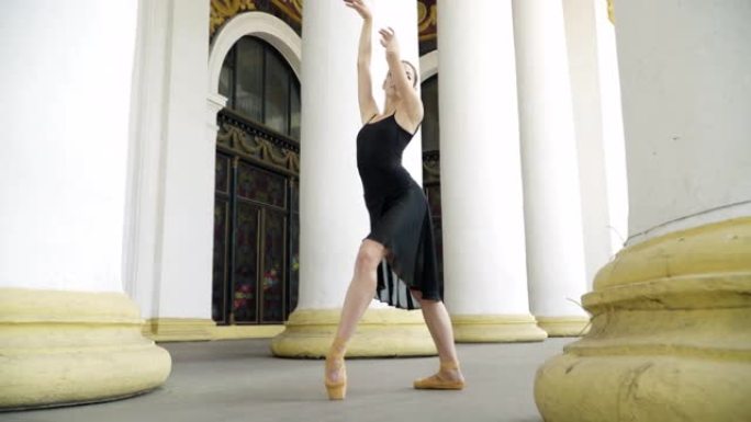 穿着黑色连衣裙的自信苗条的芭蕾舞演员在户外的柱子上缓慢移动。苗条华丽的高加索女人跳芭蕾舞的肖像。舞蹈