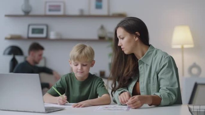 6-7岁的小儿子和年轻的妈妈家教一起学习写作。成年父母母亲教学校的孩子男孩帮助坐在家里的桌子上学习家